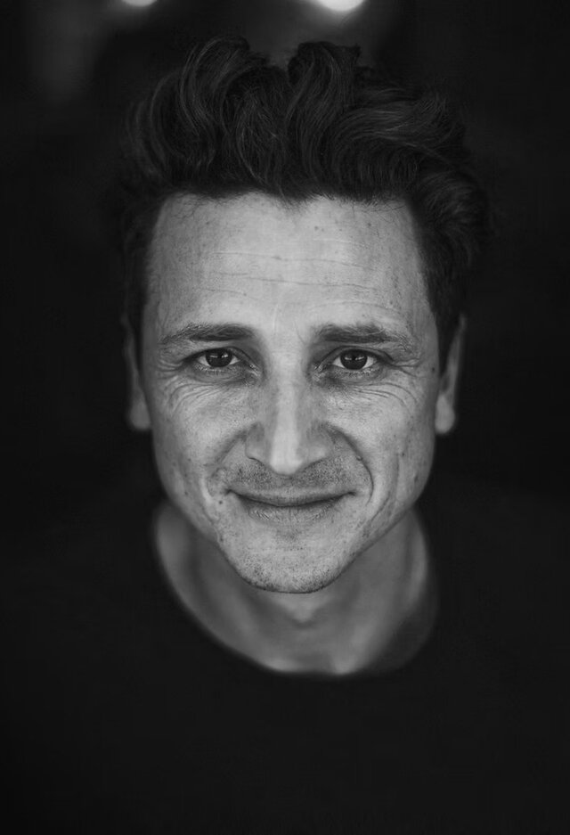 Professionelles Schwarz-Wei&szlig;-Portrait von Markus Guhl. Es zeigt ihn mit einem freundlichen, selbstbewussten L&auml;cheln und direktem Blick in die Kamera. Die Beleuchtung betont seine Gesichtsz&uuml;ge vor einem ruhigen, dunklen Hintergrund.
